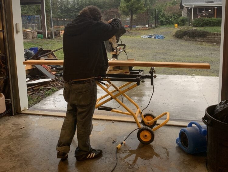 A man using a saw to cut lumber in the shelter of a shop while it rains heavily outside on the farm.