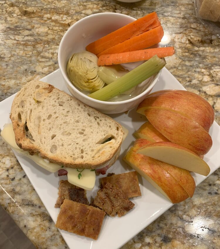 A white platter set with a sandwich, fresh apple slices, raw veggies and homemade cheese crackers.