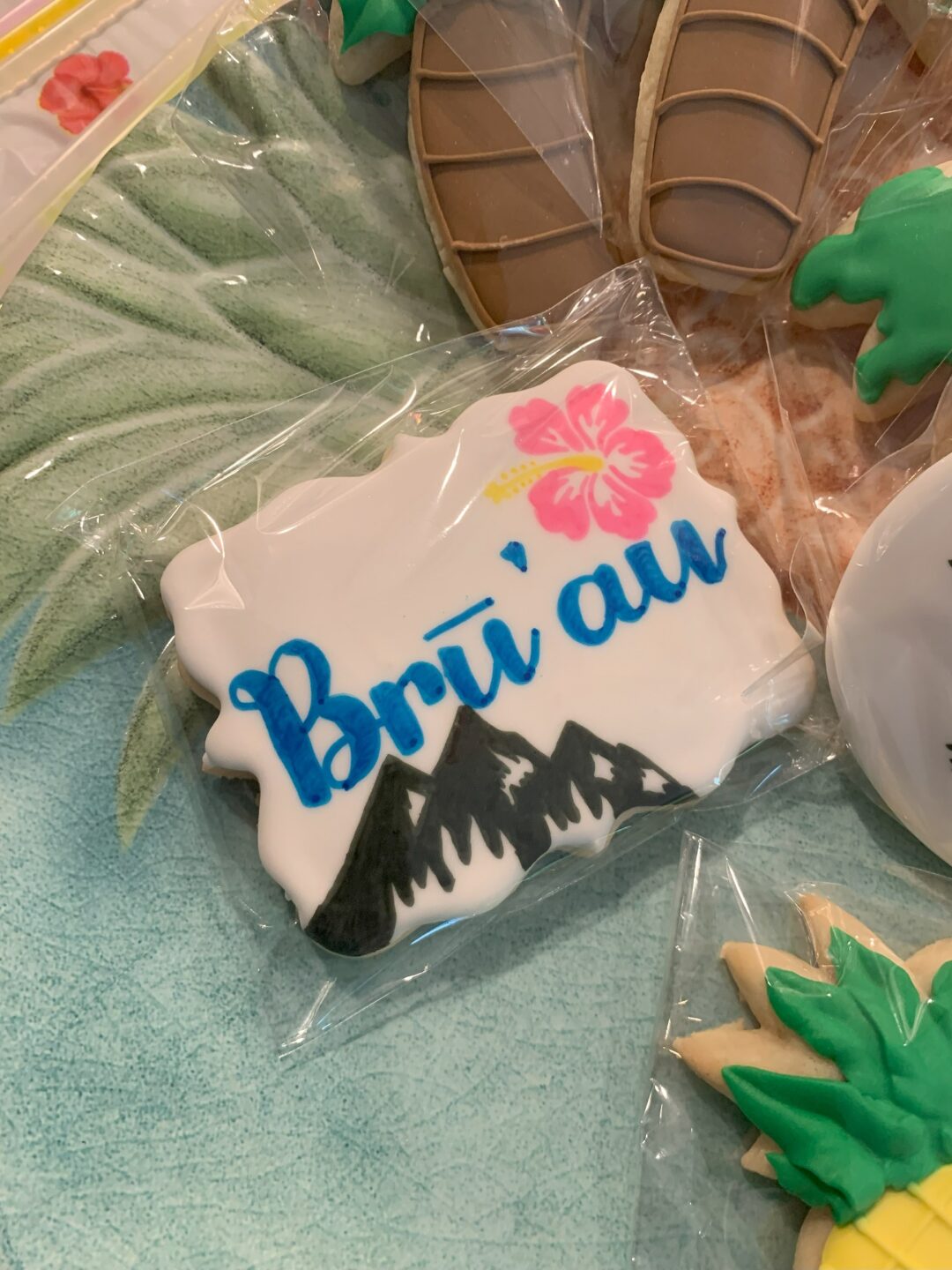 A Hawaiian-themed sugar cookie with icing decoration of a mountain, flower, and the text "Bru'au".