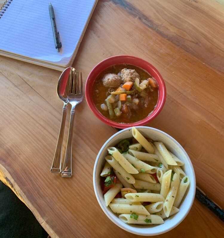 A bowl of pasta salad and bowl of soup, both sitting on a wooden table.