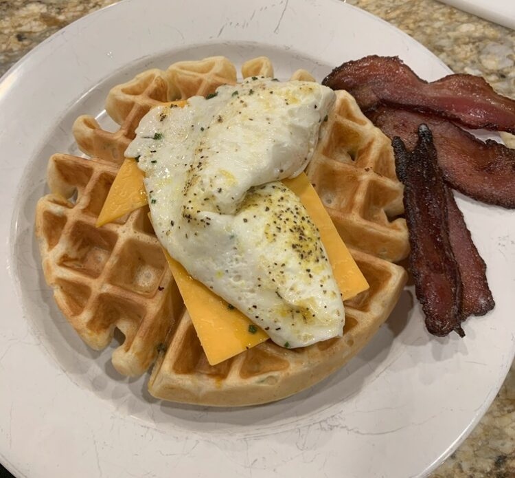 A buttermilk waffle laden with cheese and fried eggs next to a side of crispy bacon.